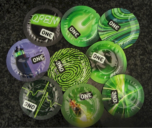 Assorted ONE Glowing Pleasures bulk condom packets in varied green and purple foil designs spread on a surface