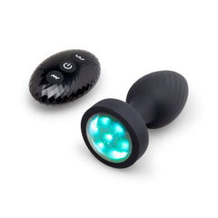 Black silicone butt plug with teal LED base lit up beside oval wireless remote