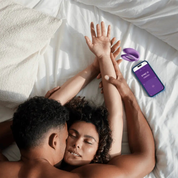 Couple on bed beside Sync O vibrator and We-Vibe app open on smartphone for remote control