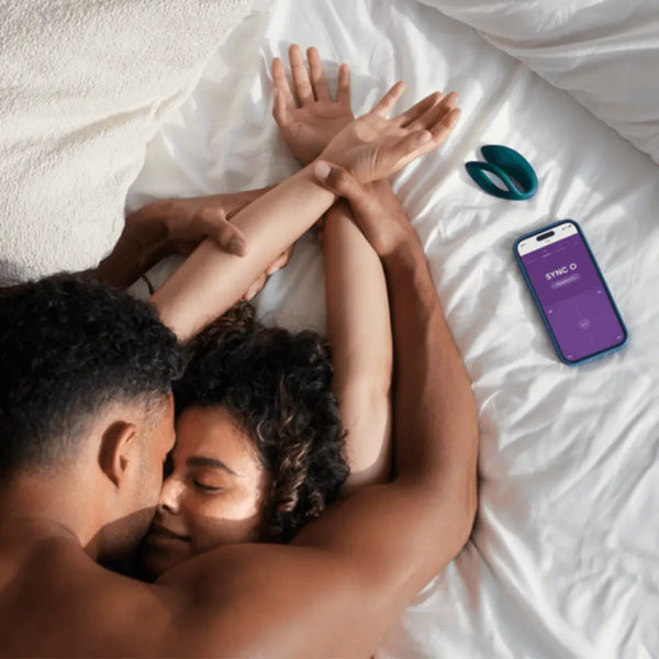 Couple on bed with Sync O vibrator and We-Vibe app open on nearby smartphone