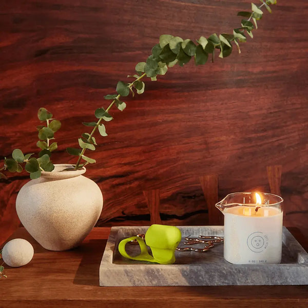 Dame Massage Oil Candle lit on marble tray beside ceramic vase with eucalyptus, warm wood backdrop