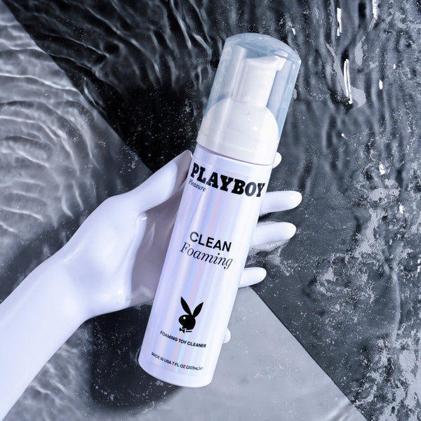Hand holding Playboy Clean Foaming Toy Cleaner bottle over flowing water background