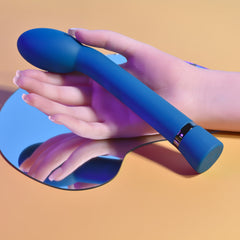 Hand resting beside blue G-spot vibrator on mirror surface, showing slim ergonomic handle