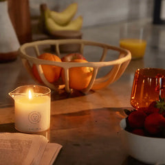 Lit Dame massage candle glowing warmly on a countertop beside fresh fruit and an open book