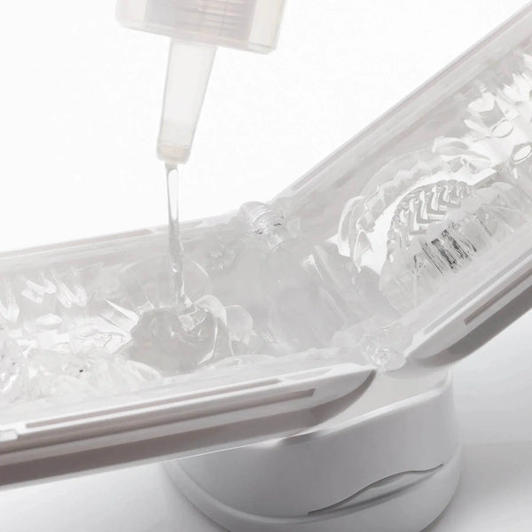 Lubricant applied from bottle into open clear textured masturbator resting on white cradle