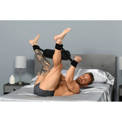 Male model on bed with ankles and wrists raised and secured together using the padded spreader bar