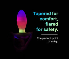 Tapered silicone anal plug glowing in rainbow LED colours held against a black background