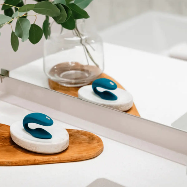 Teal We-Vibe Sync resting on a stone dish beside a bath tray with eucalyptus