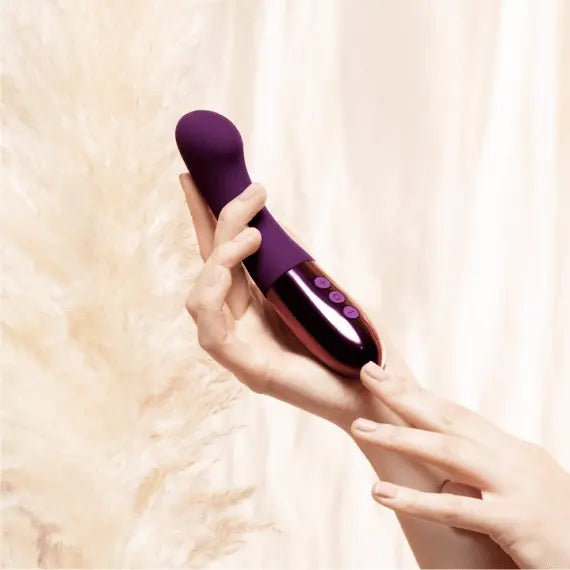 Two hands holding Le Wand Gee dark cherry vibrator against soft cream fabric backdrop