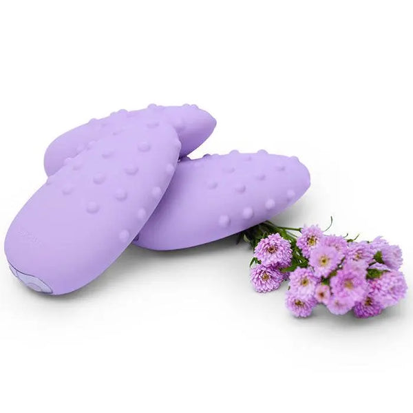 Two Mimi Deux vibrators in lavender displayed upright and flat beside purple flowers