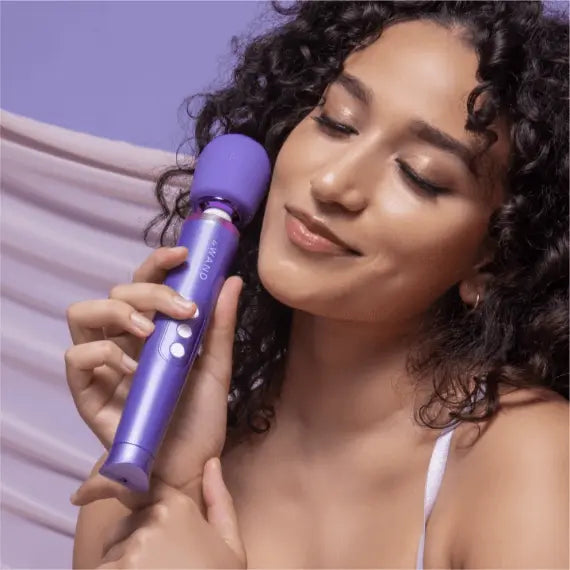 Woman with curly hair holding Le Wand Petite Massager near cheek, eyes closed, relaxed expression